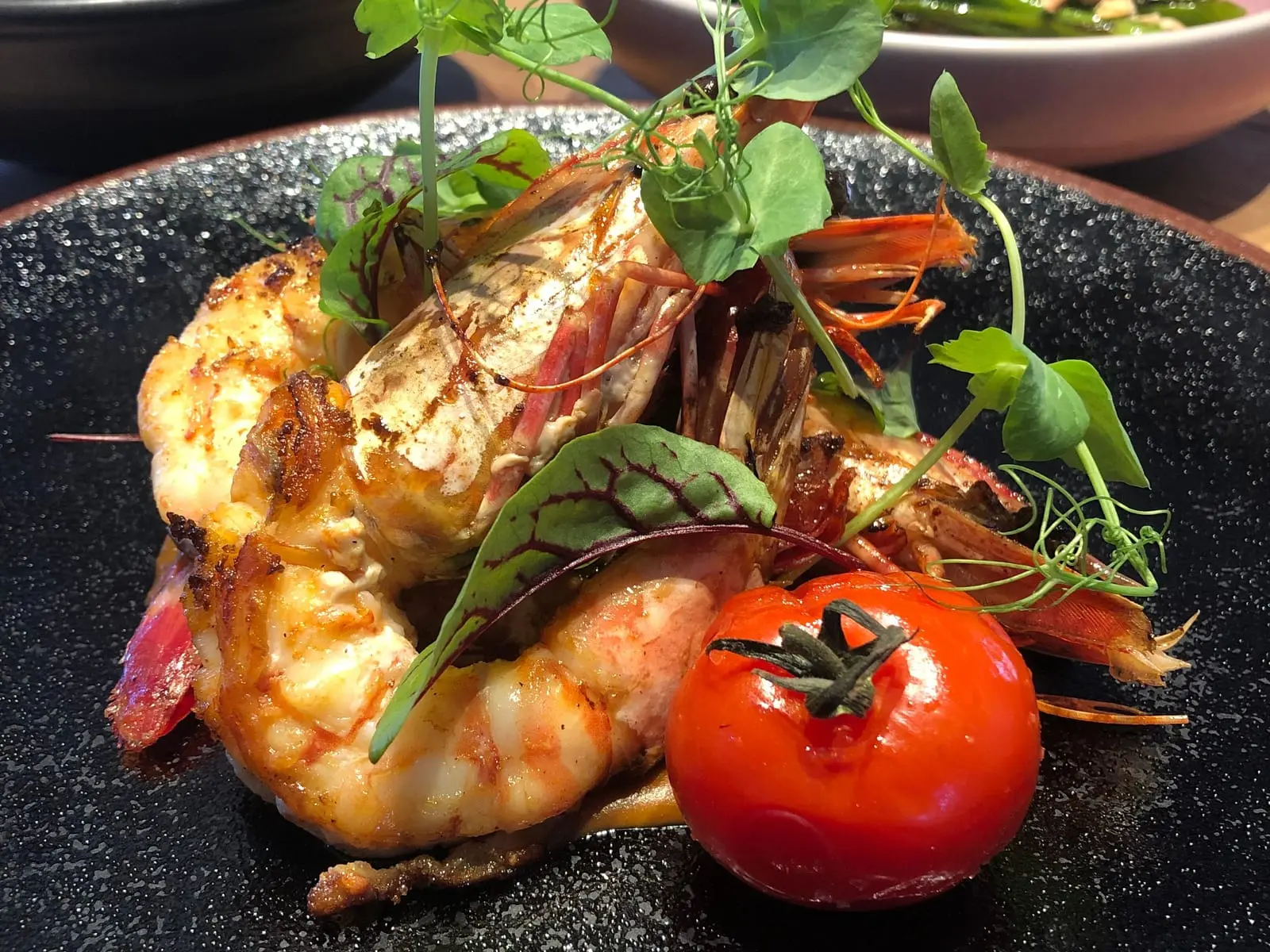Grilled king prawns with a roasted tomato and pea shoots on a dark stone plate