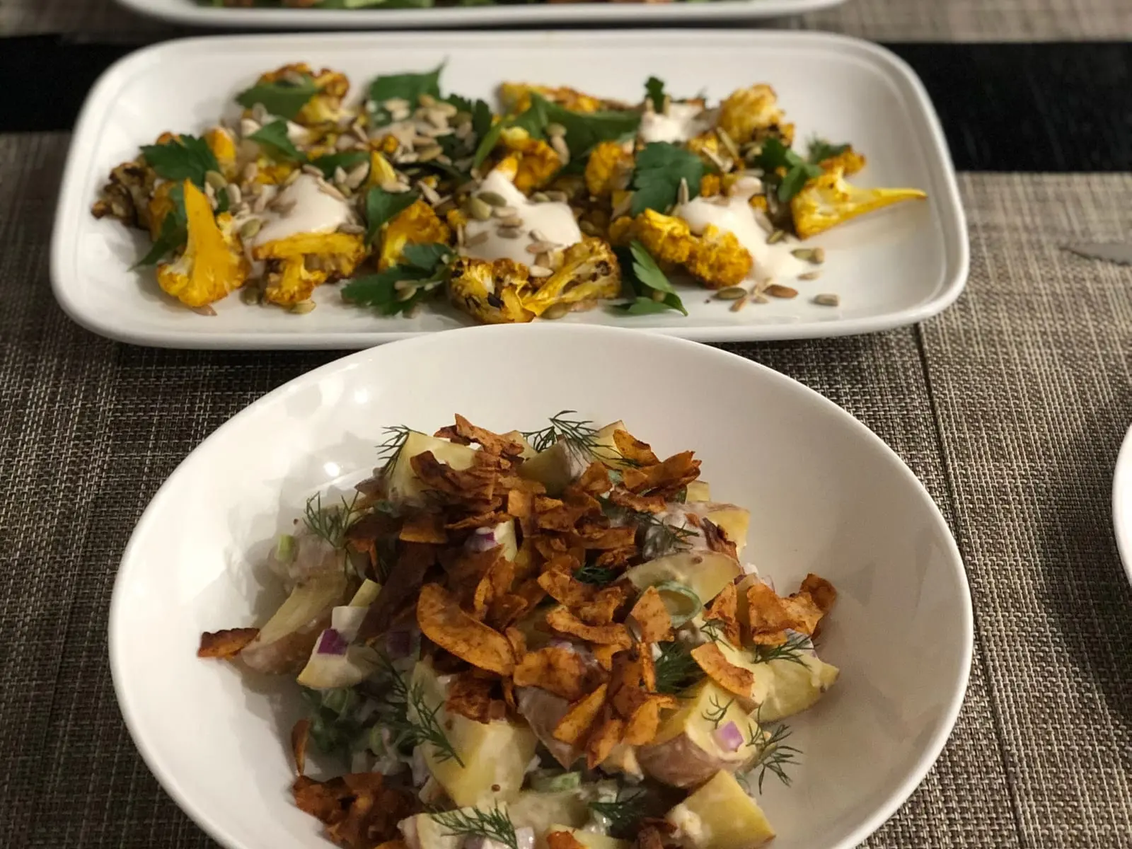Share dishes set down the middle of a dinner table: spiced roast cauliflower on a long platter and a potato salad with crisp onion and dill in a bowl