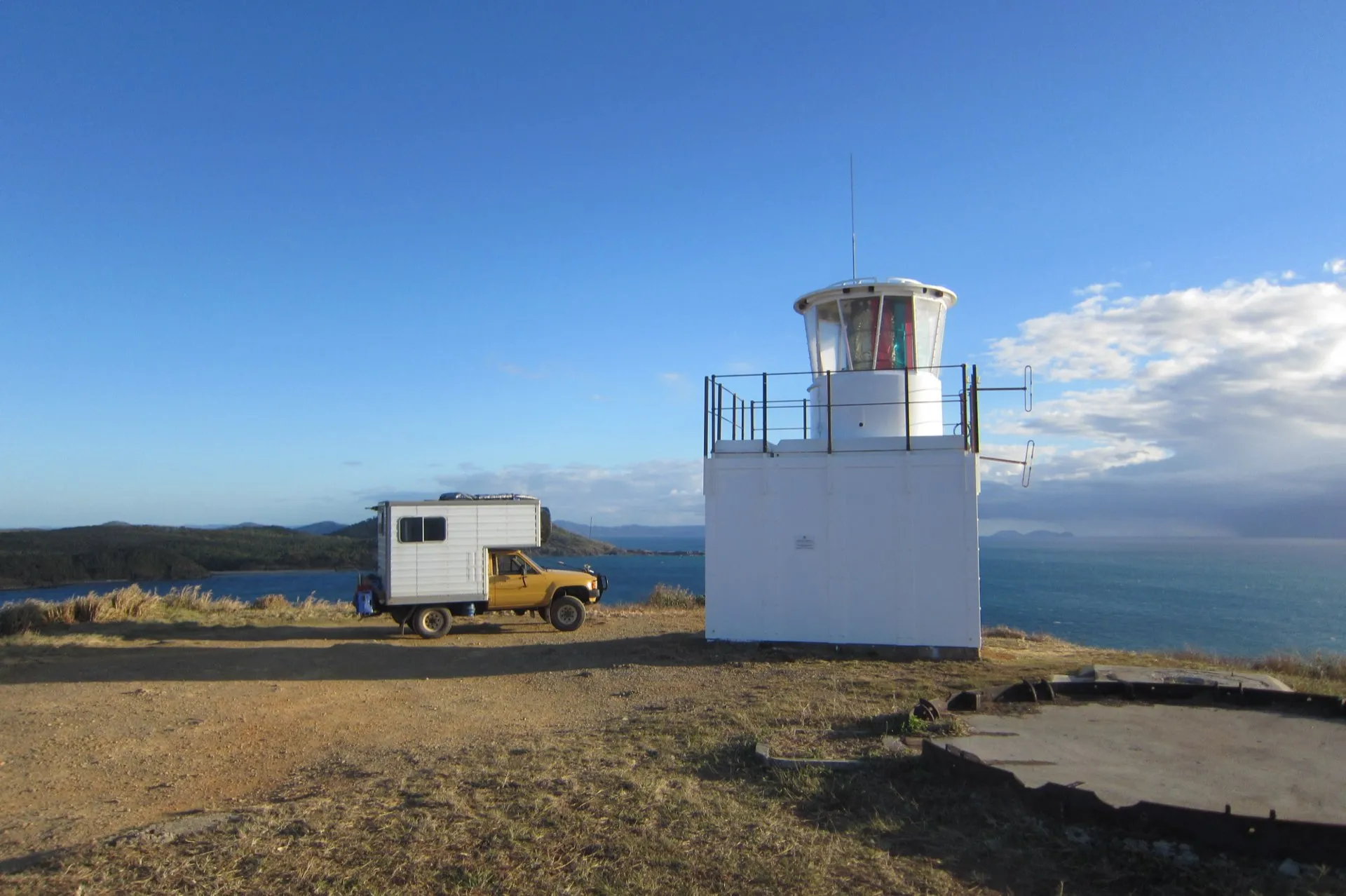 A small lighthouse and a camper alone on a remote ocean headland in late-afternoon light.