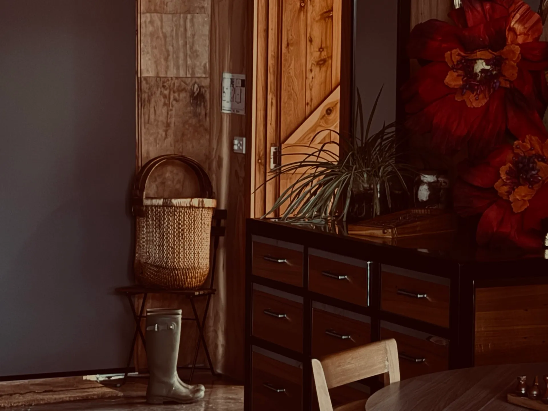A warm timber interior at dusk: a cane basket on a folding chair, gumboots by the door, a dresser with a pot plant and paper flowers.