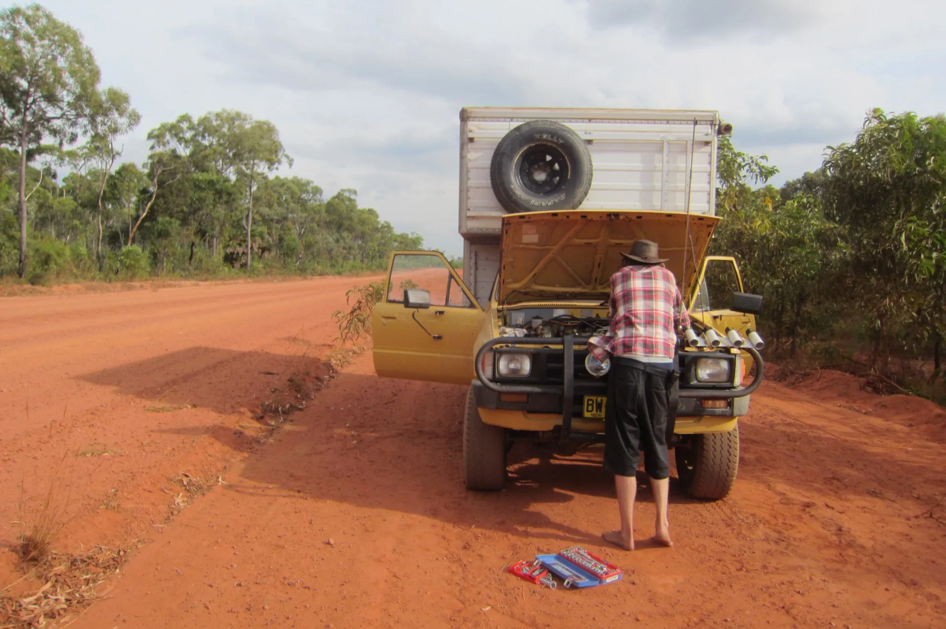 A person fixing a broken-down truck on a red-dirt road in remote country.